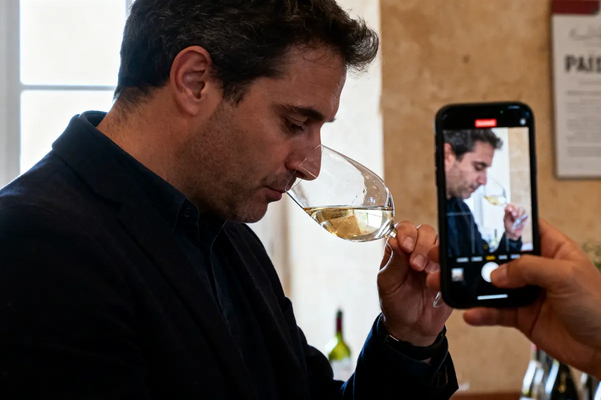 Homme dégustant du vin blanc, photographié avec un smartphone, intérieur lumineux.