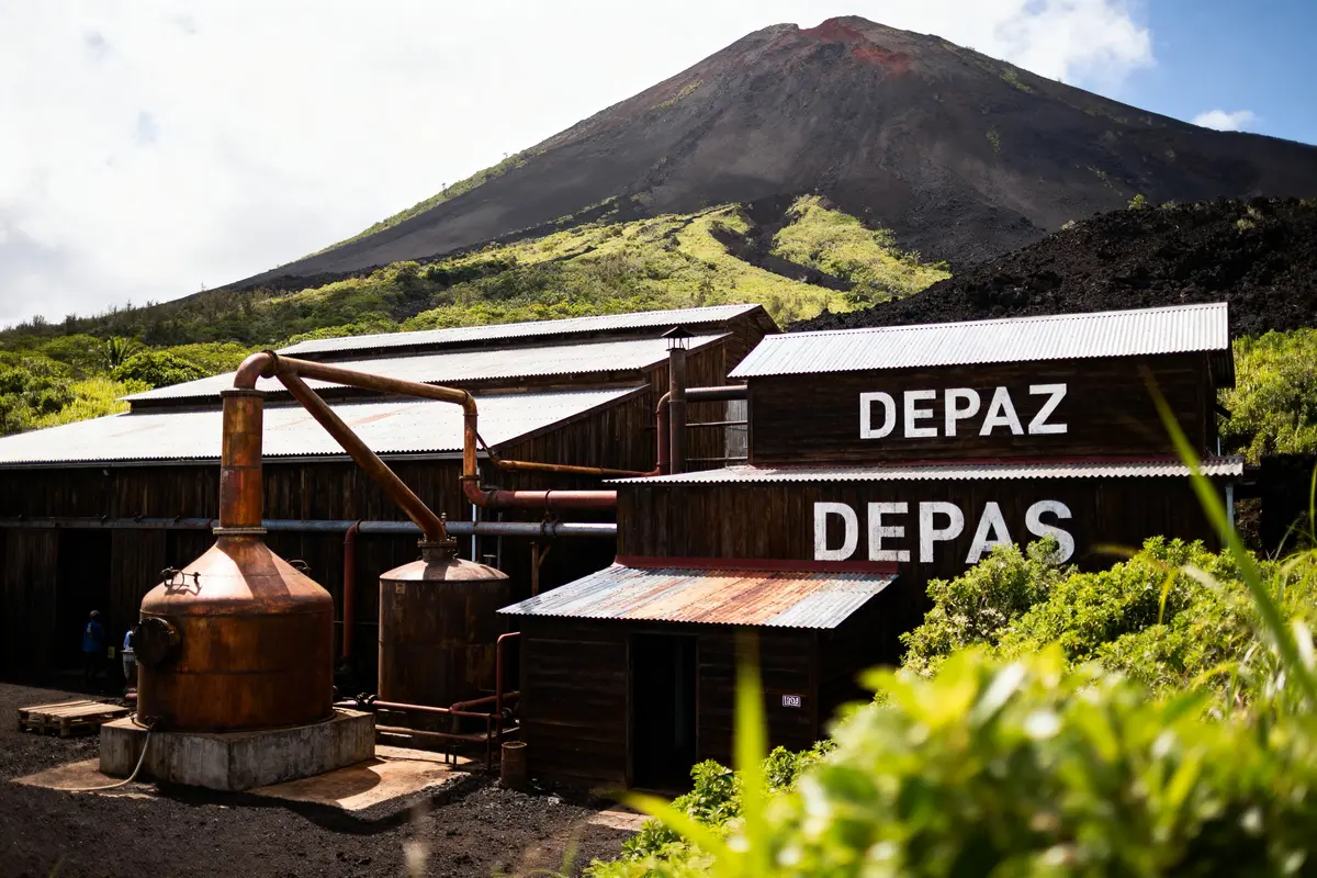 Distillerie Depaz avec alambics, en arrière-plan sommet montagne verdoyante, ciel légèrement nuageux.