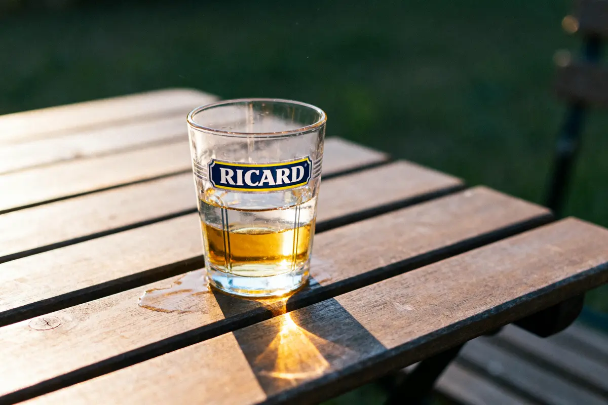 Verre de Ricard sur table en bois, éclairé par le soleil couchant, extérieur flou en arrière-plan.